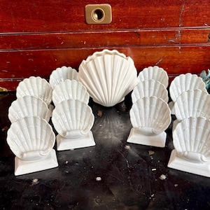 May include: A collection of white ceramic seashell-shaped dishes and a large shell-shaped bowl. The dishes are arranged on a dark surface in front of a wooden cabinet. The seashell dishes are on small rectangular bases.