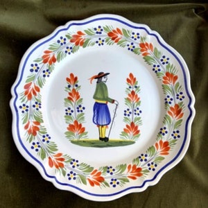 May include: A white ceramic plate with a blue and white scalloped edge. The plate features a hand-painted design of a person in traditional clothing standing in a field of red and green flowers.