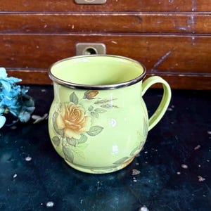 May include: A light green enamel mug with a gold rim and handle. The mug is decorated with yellow roses and green leaves. The mug is sitting on a dark surface.