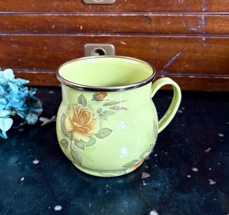 May include: A light green enamel mug with a gold rim and handle. The mug is decorated with yellow roses and green leaves. The mug is sitting on a dark surface.