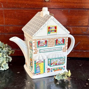 May include: A white ceramic teapot shaped like a house, with a gray roof and brick-like detailing. The teapot features the text "LA BELLA VISTA GUEST HOUSE" and illustrations of people in windows. The handle and spout are white.