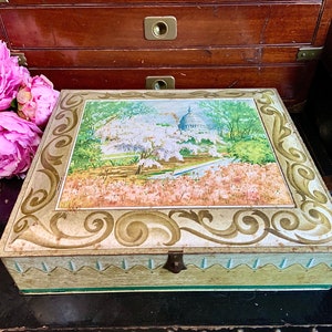 May include: A vintage tin box with a gold and green design. The lid features a painted scene of the US Capitol building surrounded by cherry blossoms.