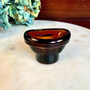May include: A small, amber-colored glass bowl with a rounded, slightly flared rim. The bowl is sitting on a white marble surface.