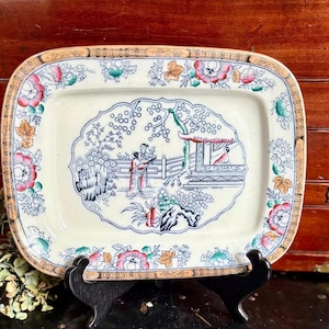 May include: Rectangular ceramic serving platter with a floral and Asian-inspired design. The platter features a central scene with figures and a pagoda, framed by floral and geometric patterns in shades of pink, green, and gold.