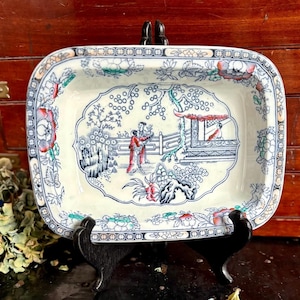 May include: Rectangular ceramic serving dish with a detailed Asian-inspired design. The dish features a central scene with figures, trees, and a pavilion, framed by floral and geometric patterns in blue, red, and green.