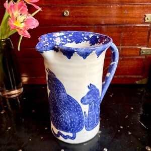 May include: White ceramic pitcher with a blue rim and handle, featuring blue illustrations of two animals. The pitcher has a spout for pouring and is likely used for serving beverages. The design is hand-painted.