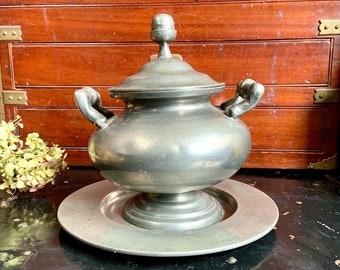 May include: A silver pewter sugar bowl with a lid and two handles, sitting on a matching silver pewter plate.