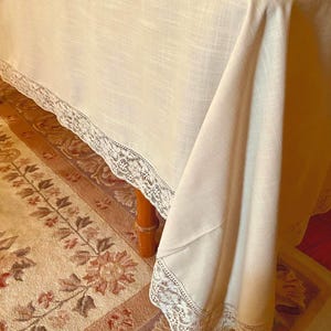 May include: A white linen tablecloth with a delicate lace trim. The tablecloth is draped over a table and hangs down to the floor. The tablecloth is on top of a patterned rug.