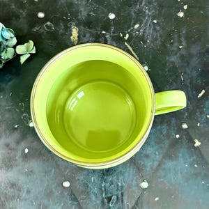 May include: Overhead view of a lime green mug with a matching handle. The interior of the mug is also lime green, and the rim has a thin gold-colored band. The mug is empty and sits on a dark, textured surface.