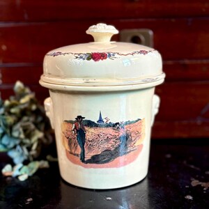 May include: Cream-colored ceramic jar with lid, featuring a pastoral scene of figures in a field with a church in the background. The lid is adorned with floral patterns. The jar has a handle on each side.