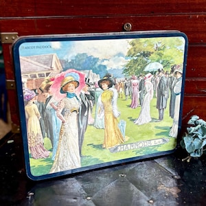 May include: Rectangular tin featuring a vintage illustration of a crowd at Ascot Paddock. The artwork depicts elegantly dressed people in hats and gowns, with the text "ASCOT PADDOCK" and "HARRODS Ld" visible. The tin has a dark blue border.