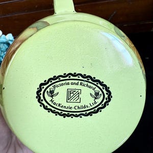 May include: A light green enamelware lid with a handle. The lid features a black and white oval logo that reads "Victoria and Richard Mackenzie-Childs, Ltd." with thistle illustrations. The lid has a floral design around the edges.