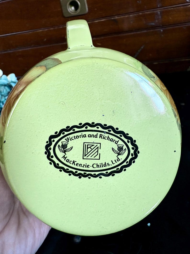 May include: A light green enamelware lid with a handle. The lid features a black and white oval logo that reads "Victoria and Richard Mackenzie-Childs, Ltd." with thistle illustrations. The lid has a floral design around the edges.