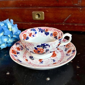 May include: A vintage teacup and saucer set. The cup and saucer feature a floral design with orange, blue, and pink accents. The teacup has a white interior and a delicate handle. The set is placed on a dark surface.