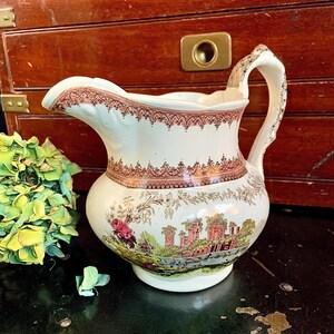 May include: A white ceramic pitcher with a brown and pink floral design. The pitcher has a handle and a spout. The design features a castle and a river.