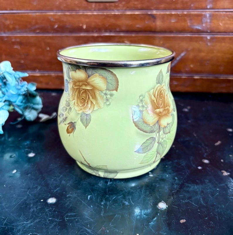 May include: A light green enamel mug with a gold rim and floral rose design. The roses are yellow and tan with green leaves. The mug has a rounded shape and is set against a dark background.