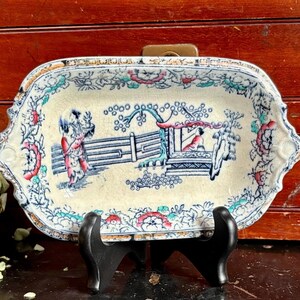 May include: Antique rectangular ceramic dish with a hand-painted Asian-inspired scene in blue, red, and green. The dish has a scalloped edge with floral and scrollwork designs. The dish is displayed on a black stand.