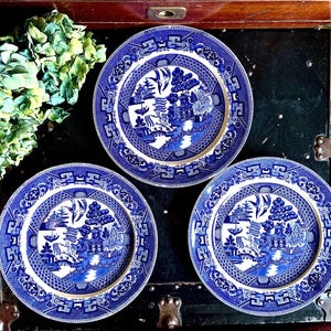 May include: Three blue and white porcelain dinner plates with a traditional willow pattern design. The plates are arranged in a triangle shape on a dark wooden surface.