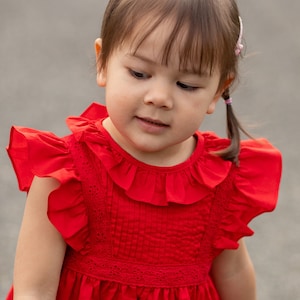 May include: A young girl wearing a red dress with a ruffled collar and sleeves. She has brown hair with pink hair clips and is looking down.