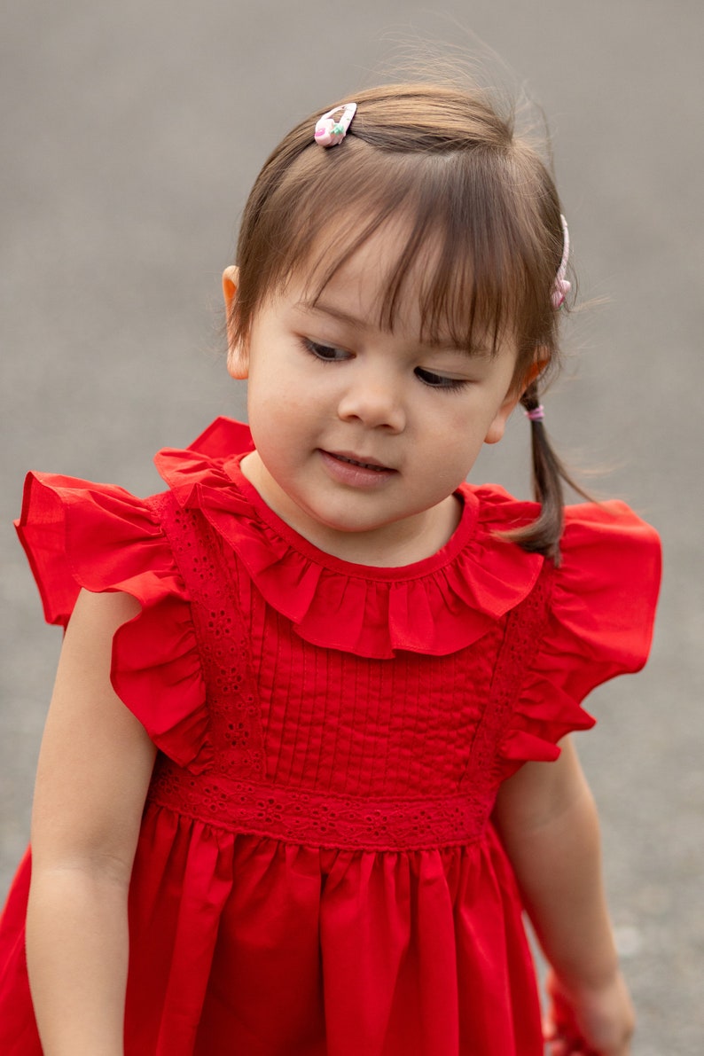May include: A young girl wearing a red dress with a ruffled collar and sleeves. She has brown hair with pink hair clips and is looking down.