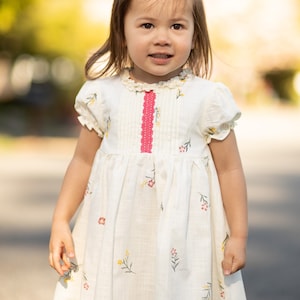 Puede incluir: Una ni&ntilde;a peque&ntilde;a con un vestido blanco con una cinta rosa y estampado floral. El vestido tiene un cuello con volantes y mangas cortas.