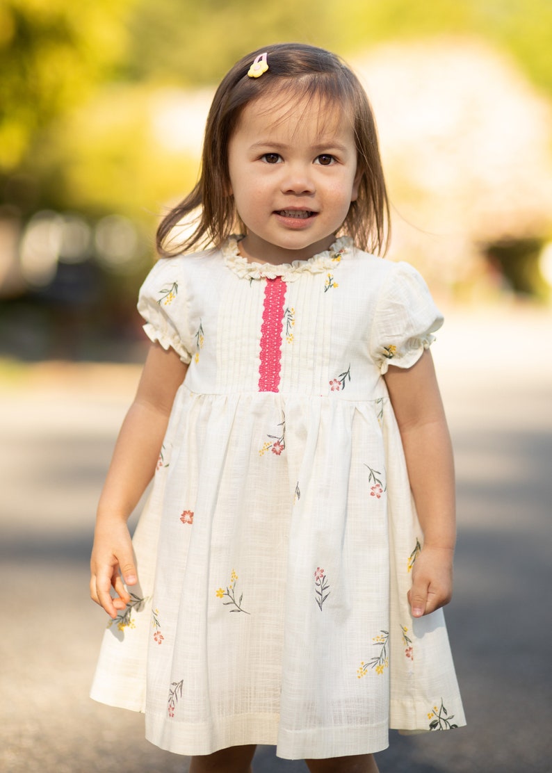 Puede incluir: Una ni&ntilde;a peque&ntilde;a con un vestido blanco con una cinta rosa y estampado floral. El vestido tiene un cuello con volantes y mangas cortas.