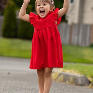 May include: A young girl in a red dress jumps in the air with her arms raised. She is wearing red shoes and has a big smile on her face.
