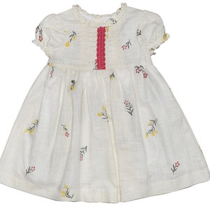 Puede incluir: Un vestido de algod&oacute;n blanco con una cinta roja y bordado floral. El vestido tiene mangas cortas y una falda fruncida.