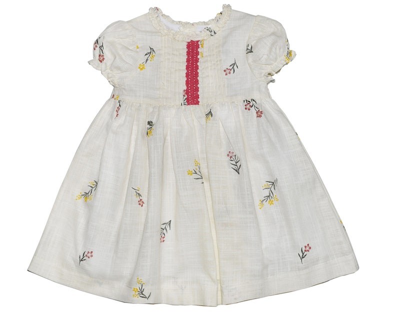 Puede incluir: Un vestido de algod&oacute;n blanco con una cinta roja y bordado floral. El vestido tiene mangas cortas y una falda fruncida.