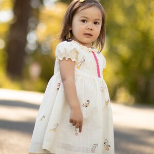 Puede incluir: Una ni&ntilde;a peque&ntilde;a con un vestido blanco con estampado floral y una cinta rosa. El vestido tiene mangas abullonadas y una falda fruncida. La ni&ntilde;a est&aacute; de pie en un camino pavimentado con un fondo borroso de &aacute;rboles.