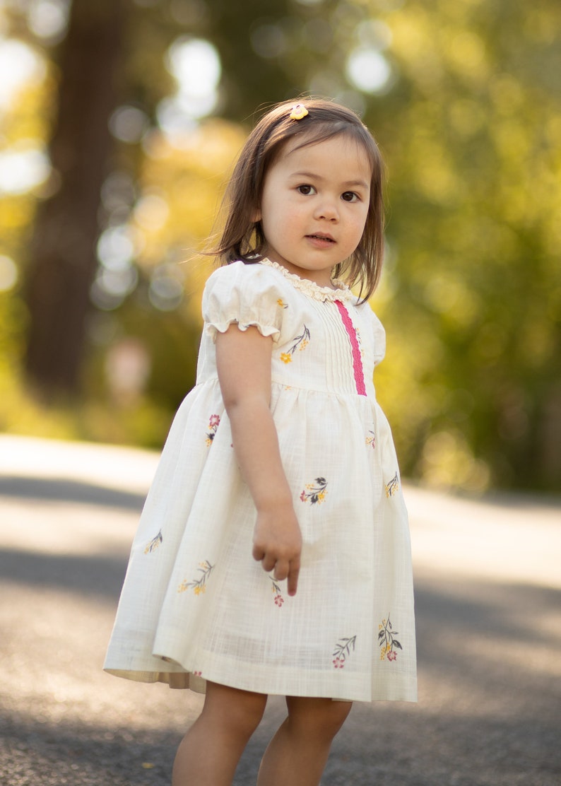 Puede incluir: Una ni&ntilde;a peque&ntilde;a con un vestido blanco con estampado floral y una cinta rosa. El vestido tiene mangas abullonadas y una falda fruncida. La ni&ntilde;a est&aacute; de pie en un camino pavimentado con un fondo borroso de &aacute;rboles.