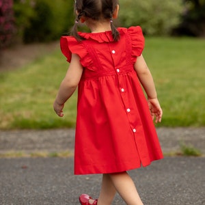 May include: A young girl wearing a red dress with white buttons and a white lace trim. Her hair is in two braids and she is wearing red shoes.