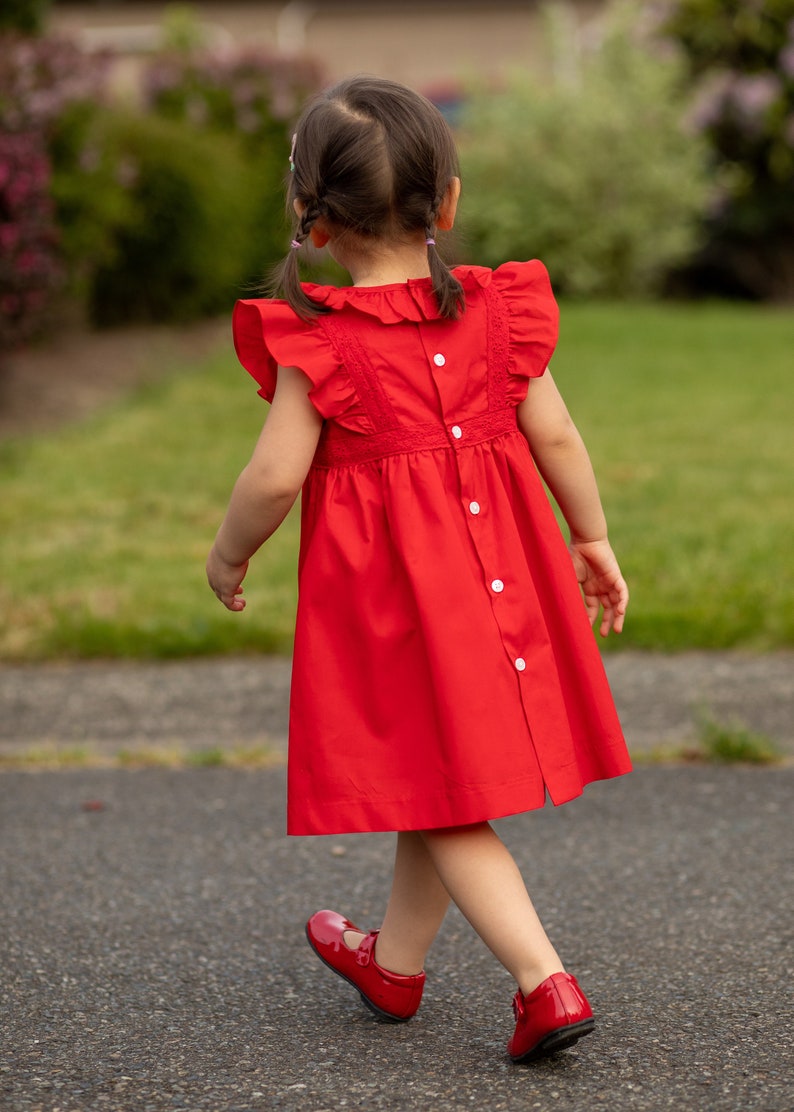 May include: A young girl wearing a red dress with white buttons and a white lace trim. Her hair is in two braids and she is wearing red shoes.