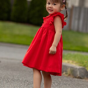 May include: A young girl wearing a red dress and red shoes. She is standing on a paved surface.