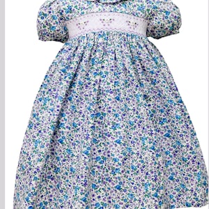 Könnte beinhalten: Ein weißes Kleid mit blauem und violettem Blumenmuster. Das Kleid hat einen Smok-Mieder und einen Rüschenkragen.