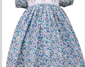 Könnte beinhalten: Ein weißes Kleid mit blauem und violettem Blumenmuster. Das Kleid hat einen Smok-Mieder und einen Rüschenkragen.