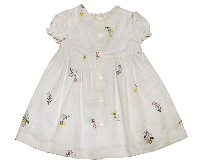 Puede incluir: Un vestido blanco con estampado floral. El vestido tiene mangas cortas y una espalda con botones.