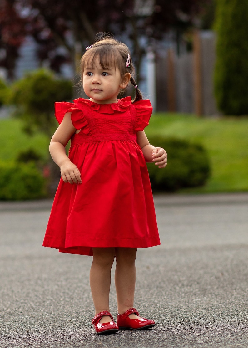 May include: A young girl wearing a red dress with ruffled sleeves and red shoes. She is standing on a paved surface.