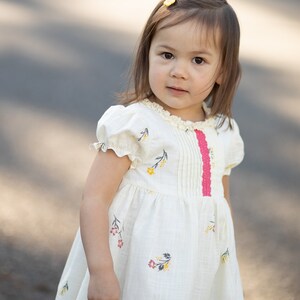 Puede incluir: Una ni&ntilde;a peque&ntilde;a con cabello casta&ntilde;o largo que lleva un vestido blanco con estampado floral y una cinta rosa. Est&aacute; de pie al aire libre en un camino pavimentado.