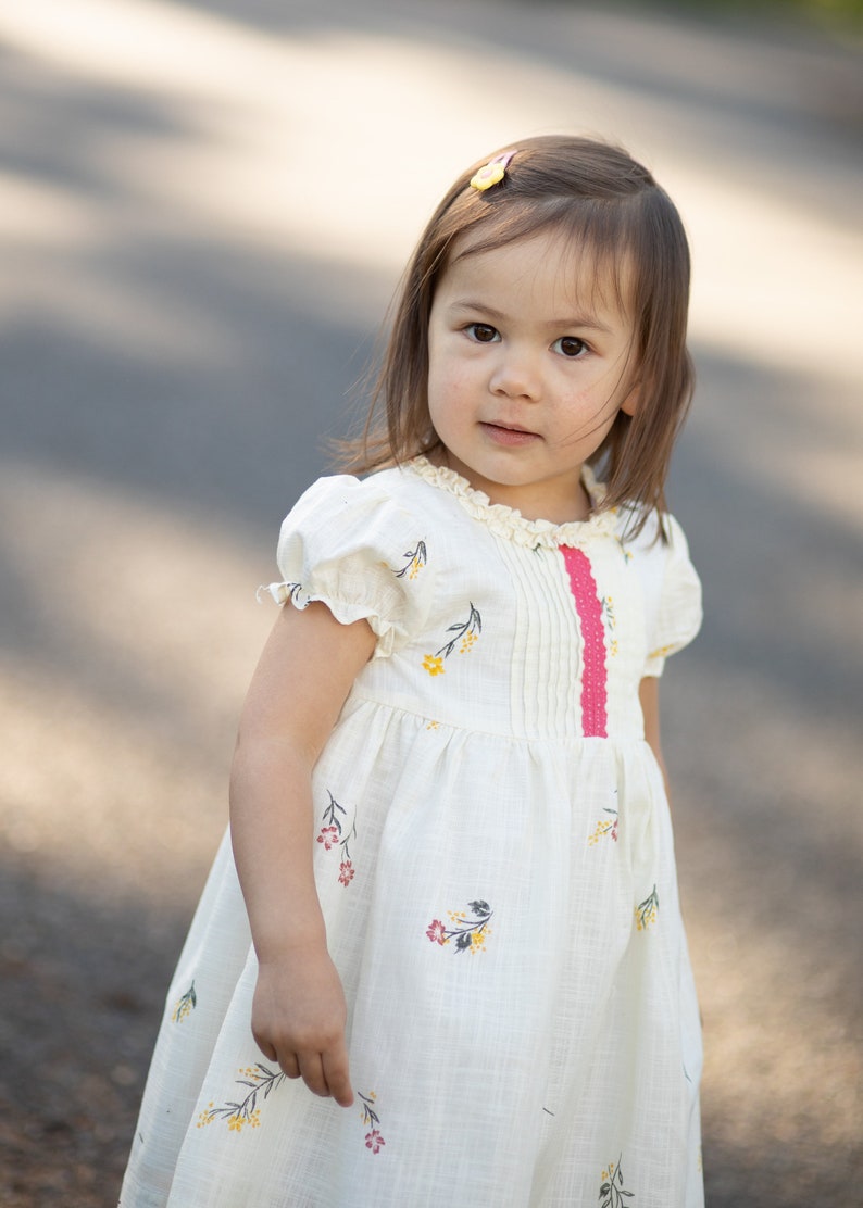 Puede incluir: Una ni&ntilde;a peque&ntilde;a con cabello casta&ntilde;o largo que lleva un vestido blanco con estampado floral y una cinta rosa. Est&aacute; de pie al aire libre en un camino pavimentado.