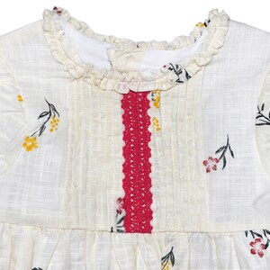 Puede incluir: Un vestido blanco con un ribete de crochet rojo en la parte delantera. El vestido tiene un cuello con volantes y un estampado floral con flores amarillas, rojas y negras.