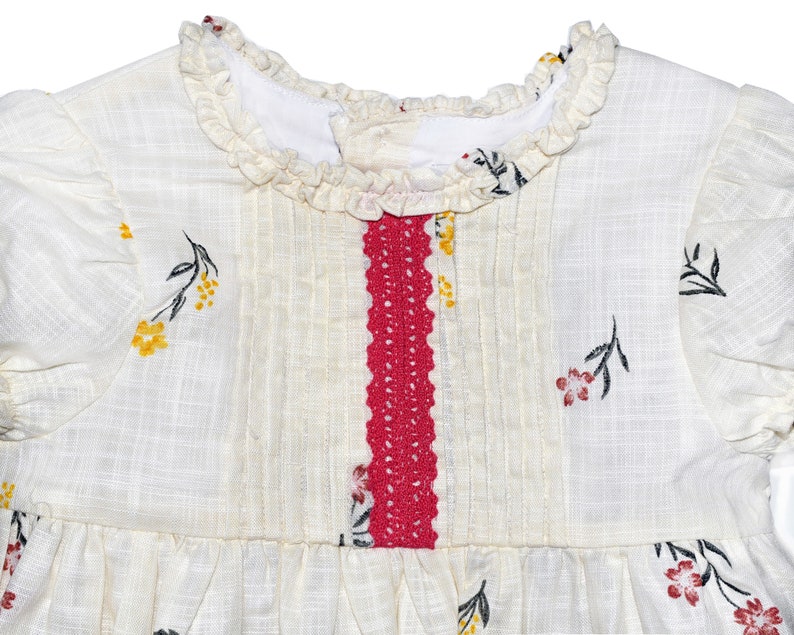 Puede incluir: Un vestido blanco con un ribete de crochet rojo en la parte delantera. El vestido tiene un cuello con volantes y un estampado floral con flores amarillas, rojas y negras.