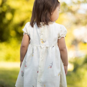Puede incluir: Una ni&ntilde;a peque&ntilde;a con un vestido blanco con estampado floral. El vestido tiene botones en la espalda y mangas cortas. La ni&ntilde;a est&aacute; de pie en un &aacute;rea cubierta de hierba con un fondo borroso.