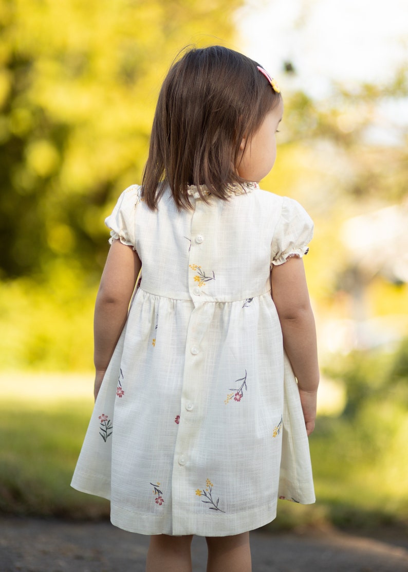 Puede incluir: Una ni&ntilde;a peque&ntilde;a con un vestido blanco con estampado floral. El vestido tiene botones en la espalda y mangas cortas. La ni&ntilde;a est&aacute; de pie en un &aacute;rea cubierta de hierba con un fondo borroso.