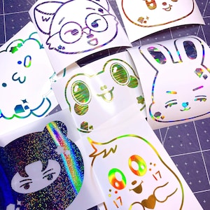 May include: A set of 8 holographic vinyl stickers featuring various cute cartoon characters. The stickers are on white paper and have a rainbow iridescent finish.