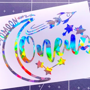 May include: A holographic vinyl decal with a crescent moon, stars, and a rocket ship. The text "To the moon and One" is written in a rainbow font.