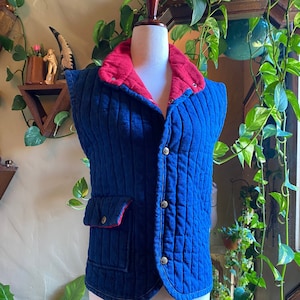 Könnte beinhalten: Eine blaue gesteppte Weste mit einem roten Cordkragen und einem einzigen Knopfverschluss. Die Weste hat eine einzelne Tasche auf der linken Seite.
