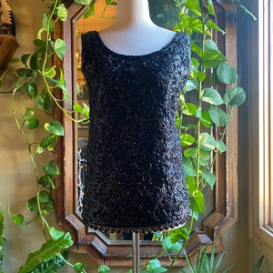 May include: A black sequined sleeveless top with a lace trim. The top is on a white mannequin and is displayed against a green plant background.