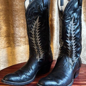 Può includere: Un paio di stivali da cowboy neri con finitura strutturata. Gli stivali presentano un design decorativo color argento sul davanti e sono appoggiati su una superficie di legno. Gli stivali hanno la punta affusolata e un tacco a blocchetto.