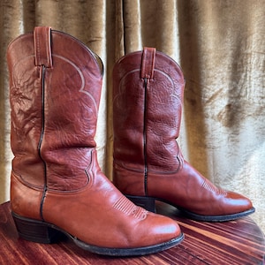 May include: A pair of reddish-brown cowboy boots. The boots have a classic design with pointed toes, decorative stitching, and pull straps. The leather appears worn, suggesting they are vintage or well-loved. The boots are set on a wooden surface.